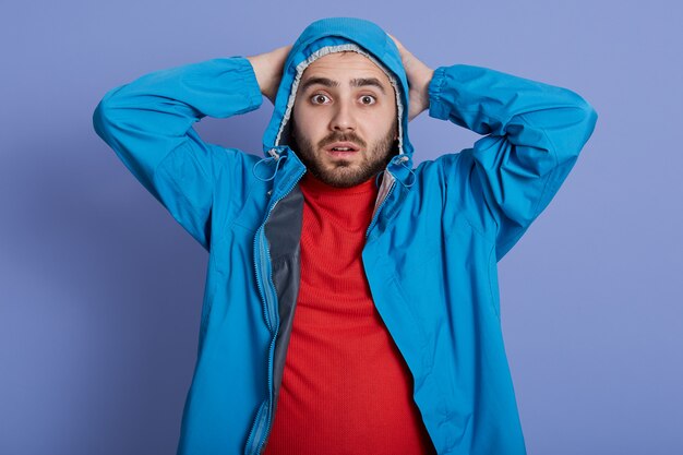 Handsome young man with wearing blue coat and red shirt standing and touching his head, looking directly at camera with big eyes