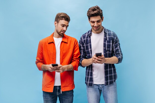 Красивые молодые люди в клетчатой рубашке и оранжевой куртке держат телефоны и позируют на изолированном синем фоне