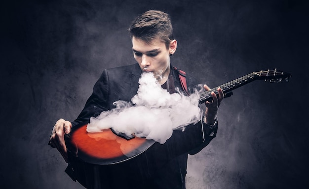 Free photo handsome young musician with stylish hair in elegant clothes exhales smoke on his acoustic guitar. isolated on a dark background.