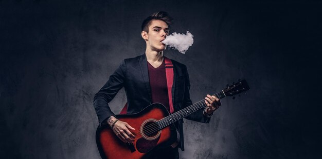 Красивый молодой музыкант со стильными волосами в элегантной одежде выдыхает дым, играя на акустической гитаре. Изолированные на темном фоне.