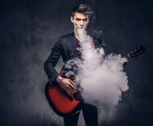 Handsome young musician with stylish hair in elegant clothes exhales smoke while playing acoustic guitar. Isolated on a dark background.