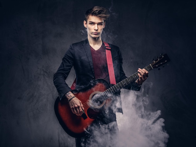 Free photo handsome young musician with stylish hair in elegant clothes exhales smoke while playing acoustic guitar. isolated on a dark background.