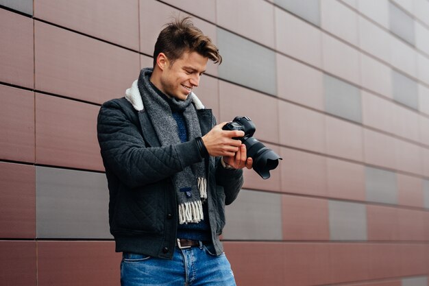 Handsome young photographer standing with camera outdoors against the building.