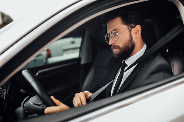 Красивый молодой серьезный бородатый водитель в полном костюме с пристегнутым ремнем безопасности за рулем автомобиля