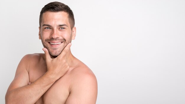 Handsome young smiling man looking at camera and keeping hand on chin while standing against white wall