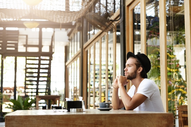 Handsome young writer with thick beard wearing black hat looking pensive and absorbed in his thoughts