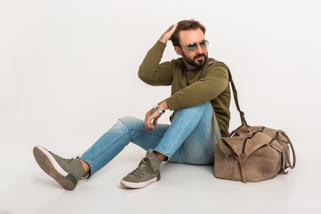 Free photo hansome bearded stylish man sitting on floor isolated dressed in sweatshirt with travel bag, wearing jeans and sunglasses