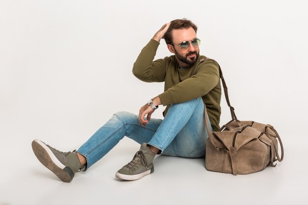 Hansome bearded stylish man sitting on floor isolated dressed in sweatshirt with travel bag, wearing jeans and sunglasses