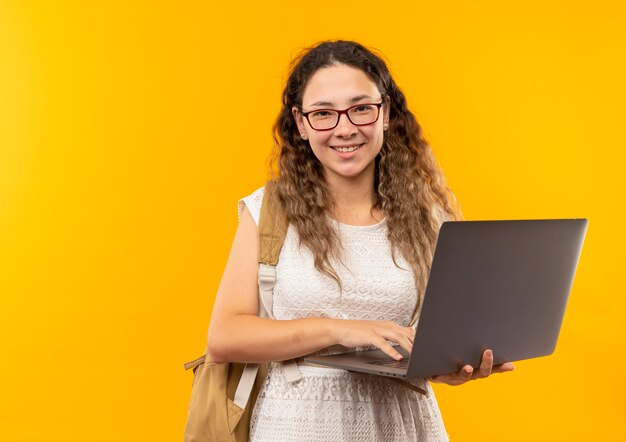 Happily smiling young pretty schoolgirl wearing glasses and back bag holding laptop isolated on yellow  with copy space