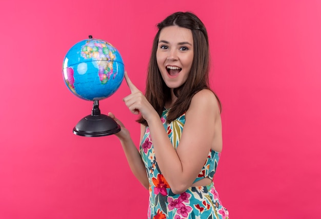 Happily smiling young traveler woman holding earth globe and pointing with finger at it on isolated pink wall