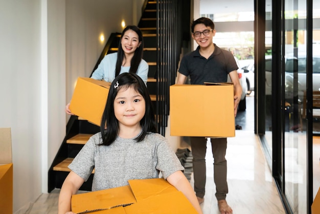 Felicità famiglia asiatica mamma papà figlia che si trasferisce nella nuova casa porta a mano una scatola di cose con un'espressione allegra e divertente