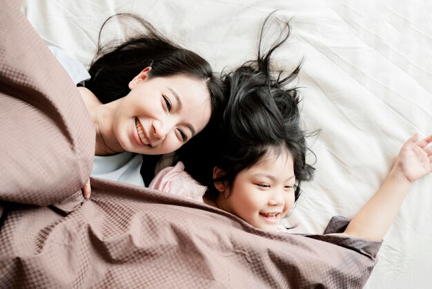 Happiness asian family moment mom and daughter play and laydown with blanket on white bed home interior background