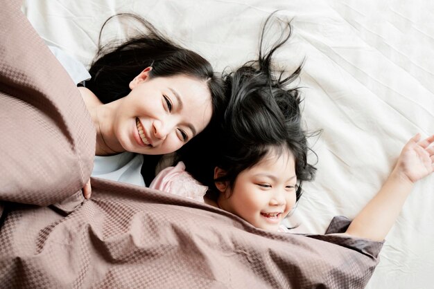 Счастье азиатской семьи момент, когда мама и дочь играют и ложатся с одеялом на фоне интерьера дома белой кровати