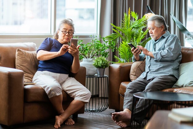 행복 아시아 은퇴한 부부는 거실 홈 인테리어 배경에서 소파에서 온라인으로 스마트폰 모바일 게임 및 경쟁 게임을 즐깁니다.