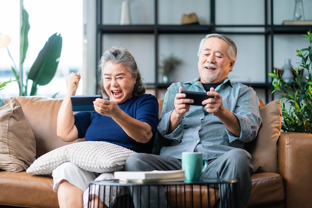 행복 아시아 은퇴한 부부는 소파 거실 홈 인테리어 배경에서 함께 온라인으로 스마트폰 모바일 게임 및 경쟁 게임을 즐깁니다.