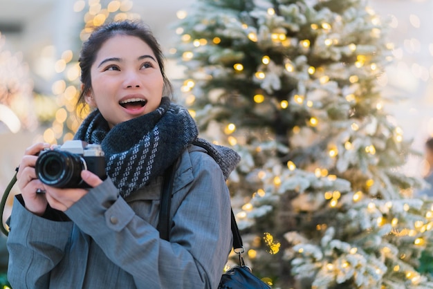 무료 사진 아름다운 아시아 여성의 손을 잡고 있는 카메라는 조명 장식과 황금빛 흐림 배경이 있는 크리스마스 트리를 즐깁니다.