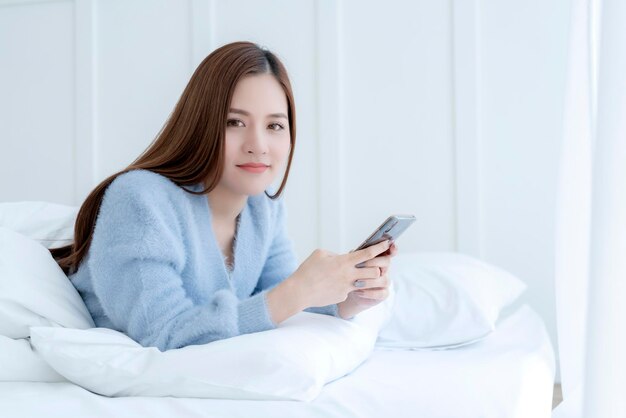 幸福美しいアジアの女性の笑顔の朝の目覚め白いベッドの柔らかい枕と手持ちスマートフォンの寝室の背景