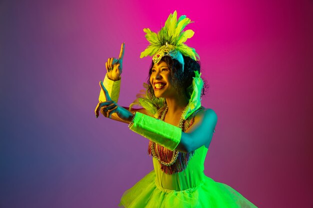 Happiness. Beautiful young woman in carnival, stylish masquerade costume with feathers dancing on gradient background in neon. Concept of holidays celebration, festive time, dance, party, having fun.