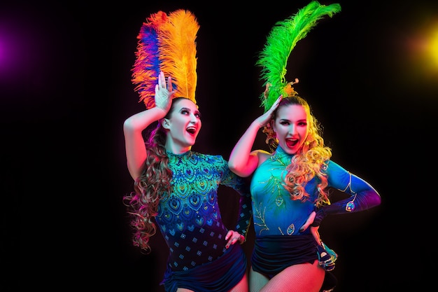 Happiness. Beautiful young women in carnival, stylish masquerade costume with feathers on black background in neon light. Copyspace for ad. Holidays celebration, dancing, fashion. Festive time, party.