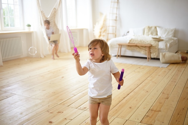 Happiness and carefree childhood concept. Indoor portrait of adorable happy female child in t-shirt and shorts standing in the middle of modern living room, having fun, blowing soap bubbles,