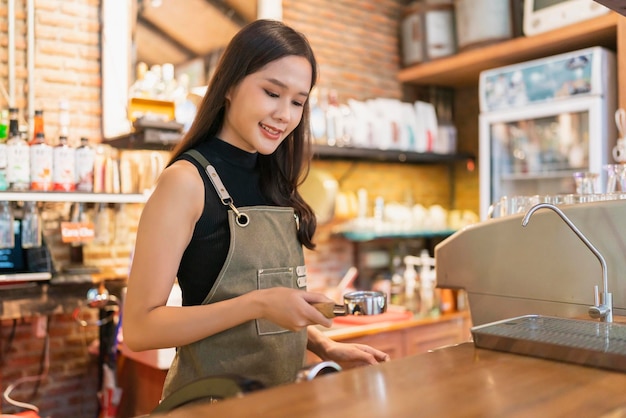 カフェショップ店でコーヒーマシンでコーヒーを作るコーヒーショップで働く中国の女性バリスタで働く制服中小企業コーヒーショップアジア女性オーナーの幸せ陽気なアジアの女性