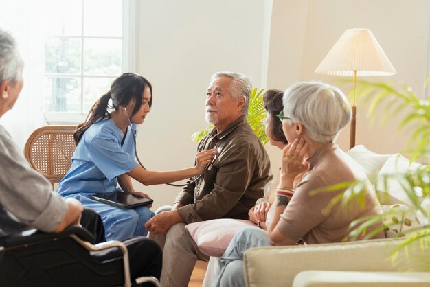 Happiness Cheerful elderly woman and men talking with female caregiver nurse doctor having health checking consult at living areaCaretakers with senior couple sitting in living room at nursing home