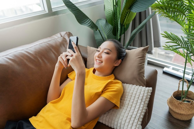 Felicità allegra sorridente giovane donna asiatica adulta indossa panno giallo conversazione a mano sullo smartphone sul divano a casa quarantena remota periodo di blocco sfondo interno domestico