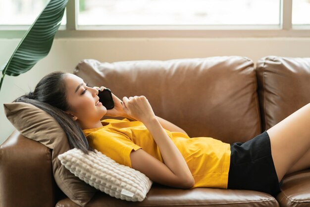 Happiness cheerful smiling young adult asian female woman wear yellow cloth hand conversation on smartphone on sofa at home remote quarantine lock down period home interior background