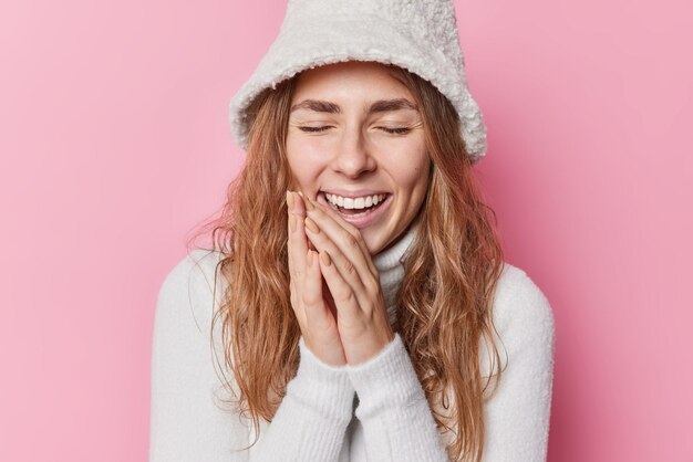 Happiness concept Sincere European woman keeps hands together laughs loudly with closed eyes has upbeat mood wears white panama and jumper with collar recalls funny story happened with her