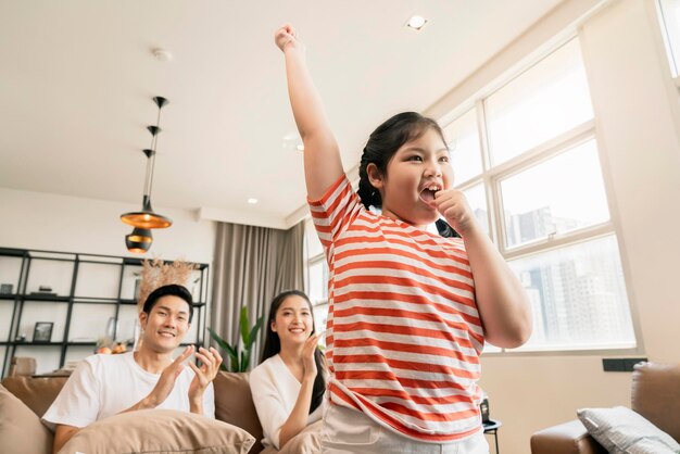 幸せの喜びアジアの素敵な家族は、リビングルームのテレビの前で小さな娘の女の子の歌とダンスの練習を見て元気づけて楽しんでいます