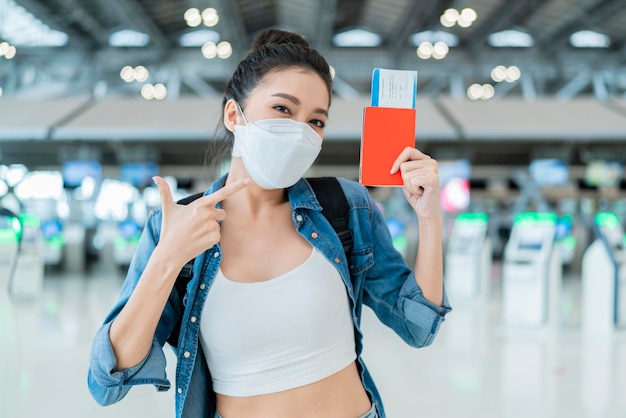 Счастье, улыбающаяся азиатская взрослая женщина в маске для лица, жест рукой, показывающая паспорт и смартфон с пустым экраном в терминале аэропорта