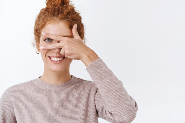 Happiness wellbeing and lifestyle concept Gorgeous feminine lovely redhead woman with curly hair combed in bun wear stylish blouse showing peace sign as cover face with hand smiling