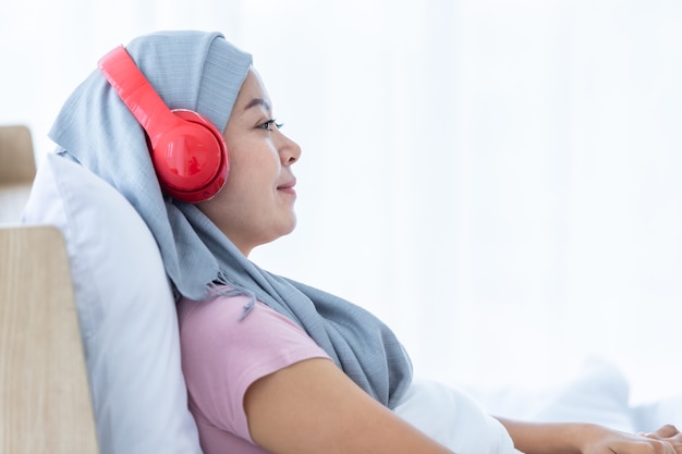 헤드폰에 머리 스카프를 두른 분홍색 리본이 달린 행복한 아시아 여성 유방암 환자가 음악을 듣고 있습니다. 화학요법 치료 후 집 침실에 앉아