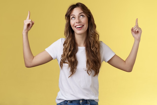 Felice ammirando carino europeo ragazza dai capelli ricci taglio di capelli lungo look rivolto verso l'alto impressionato divertito sorridente ampiamente soddisfacente felice stand t-shirt bianca sfondo giallo reagire con gioia buon promo