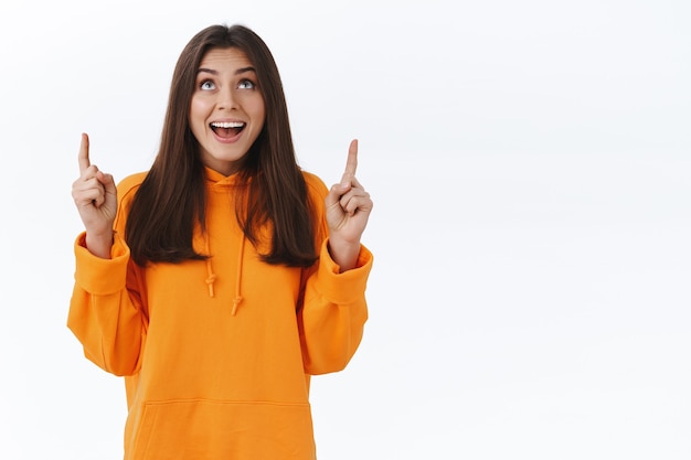 Happy adorable brunette woman in orange hoodie pointing and looking up with cheerful expression, found something really awesome and entertaining, standing enthusiastic white wall