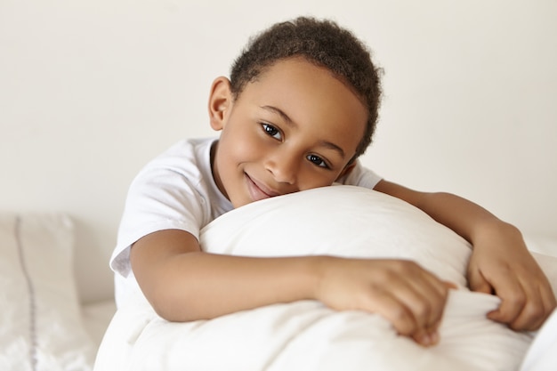 Happy adorable dark skinned boy of African origin relaxing in bed at weekend after awakening