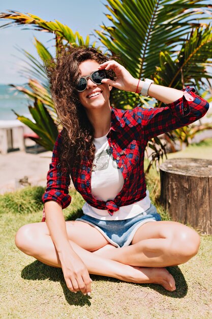 Happy adorable stylish pretty woman with curly dark hair dressed white t-shirt and denim shorts sitting on green land by palm leaves in sunshine, vacation, travel, spa resort