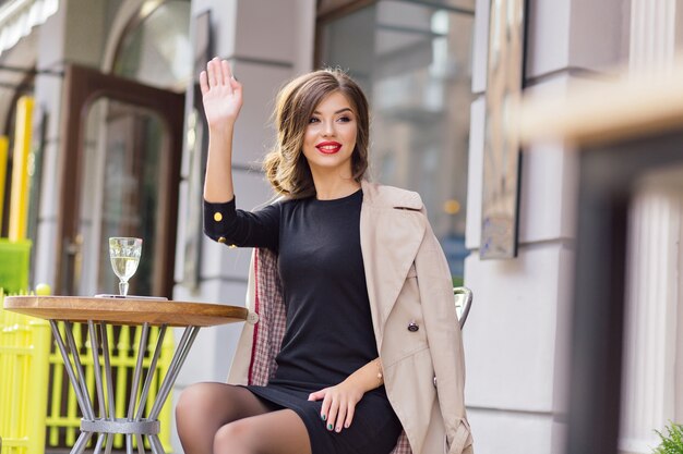 Happy adorable woman waving to friend while sitting in summer cafeteria with a glass of wine