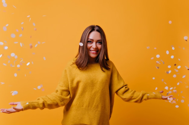 Happy adorable woman in yellow sweater laughing on bright wall. Indoor portrait of appealing girl posing with confetti.