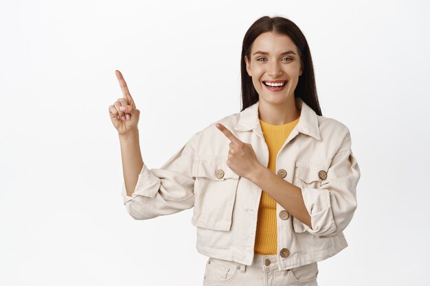 happy adult brunette woman pointing fingers at sale on upper left corner, showing advertisement, banner or logo of company, standing on white.