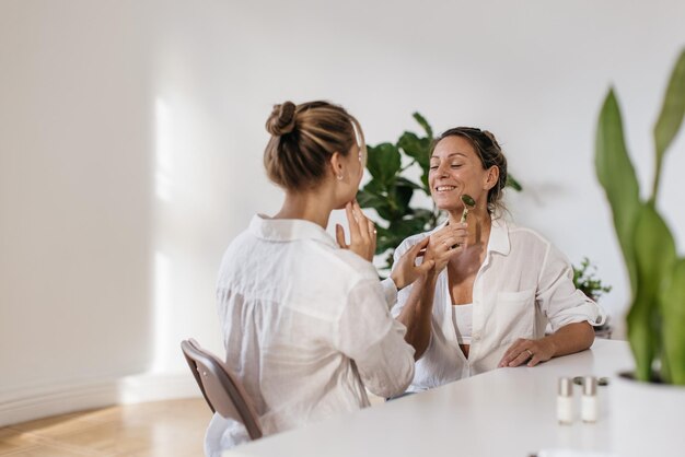 Happy adult caucasian lady uses roller massager while sitting next to blonde woman at table indoors. Skin care concept