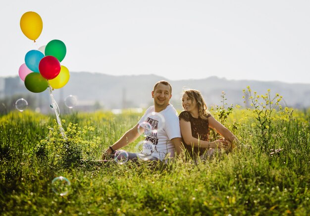Happy adult couple has fun on a green field sitting with colorful balloons 