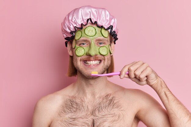 幸せな大人の男性は、顔にキュウリのスライスが付いた緑の栄養マスクを適用します。歯ブラシは、半分裸の屋内で歯を磨き、朝の毎日の衛生ルーチンを持っています。