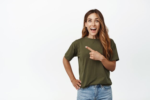 Happy adult woman pointing finger left, gasping and smiling excited, showing advertisement, making big announcement, standing against white background.