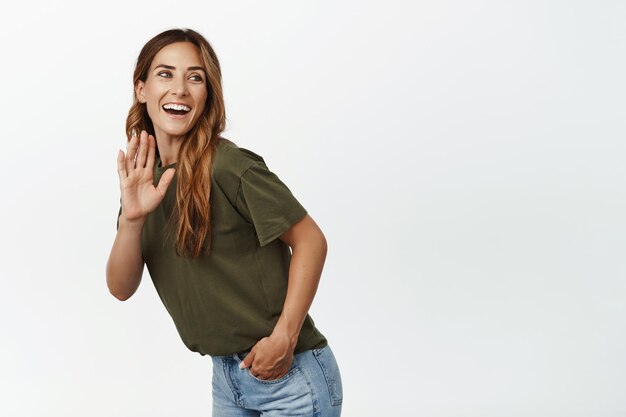 Happy adult woman waving hand and smiling, greeting gesture, standing in tshirt against white background