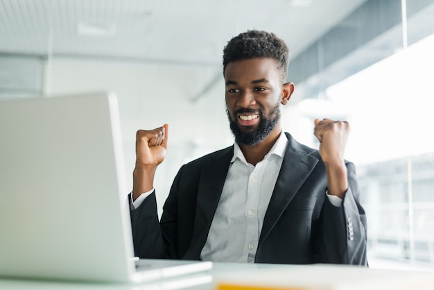 Happy african-american businessman in suit looking at laptop excited by good news online. Black man winner sitting at office desk achieved goal raising hands celebrating business success win result