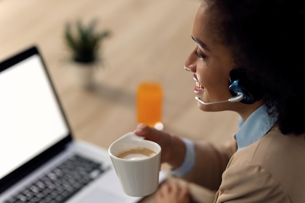 Caffè bevente della donna di affari dell'afroamericano felice durante la teleconferenza sopra il computer portatile