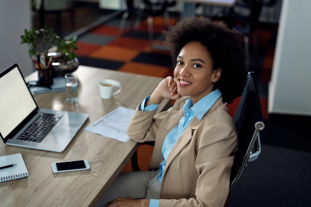 Donna d'affari afroamericana felice che lavora al computer portatile in ufficio e che guarda l'obbiettivo