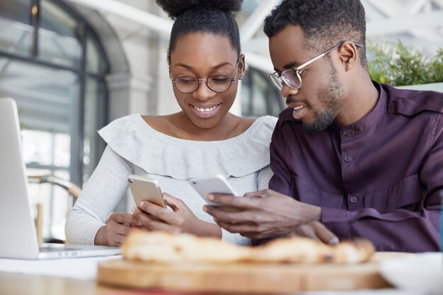 起業家の幸せなアフリカ系アメリカ人カップルがポータブルラップトップコンピューターで新しいビジネス戦略を開発し、インターネットで情報を閲覧するために携帯電話を使用する