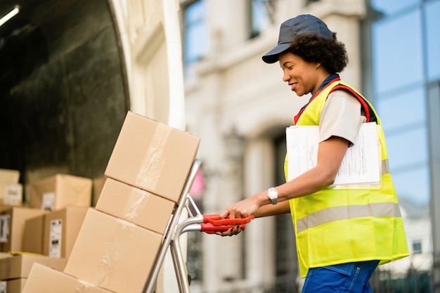 Happy African American deliverer with hand truck unloading packages from a van